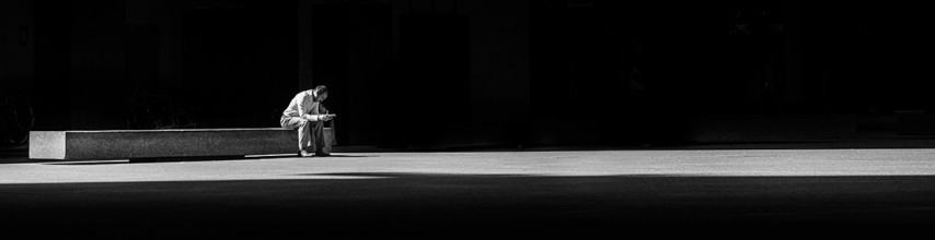 A person sitting alone on a long concrete bench. The person is in grey, the seat is grey and the background is black