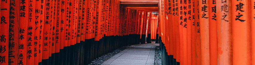 Red flags along a walkway. They have either Chinese characters of Japanese kanji written in black on the flags. The flags have painted black bottoms and are alongside a grey stone and concrete path