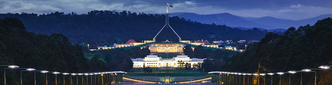 A picture of Parliament House. It is a white building with a spire on the top. There are lights lighting up the building. There is a green lawn and dark blue sky with a darker blue outline of mountains and trees